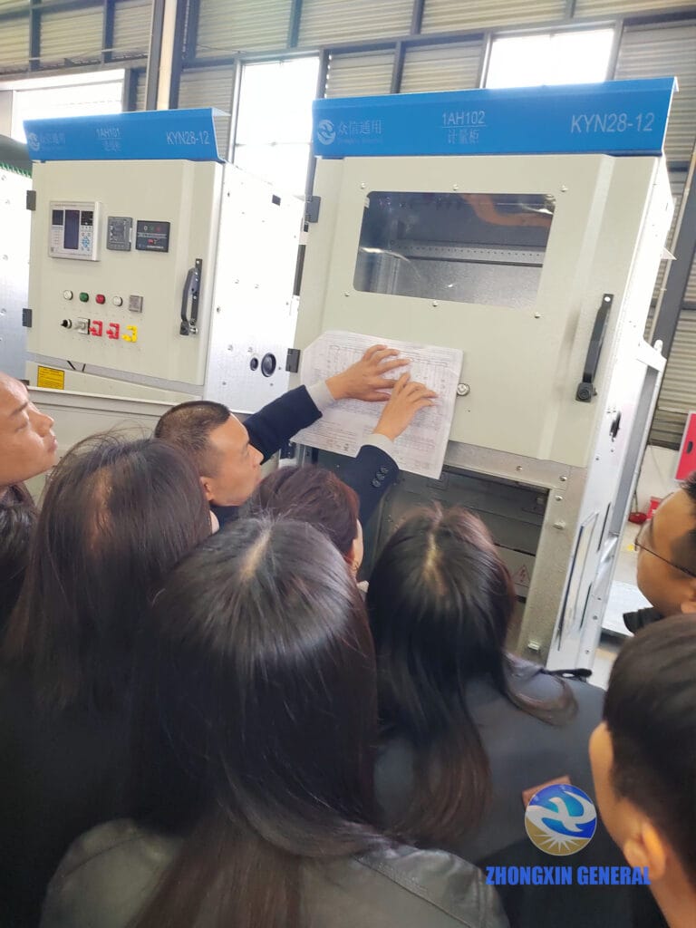 He Dongqi demonstrating technical drawings against a KYN28-12 medium-voltage switchgear cabinet in Zhongxin General's production workshop, with sales team members crowded around to observe.