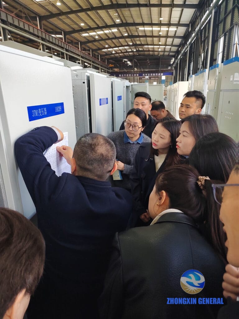 He Dongqi, Director of Complete Equipment Engineering, pointing to a transformer nameplate on a '3TM1 Transformer 1AH209' unit in the Zhongxin General production workshop, with sales staff gathered closely around him.